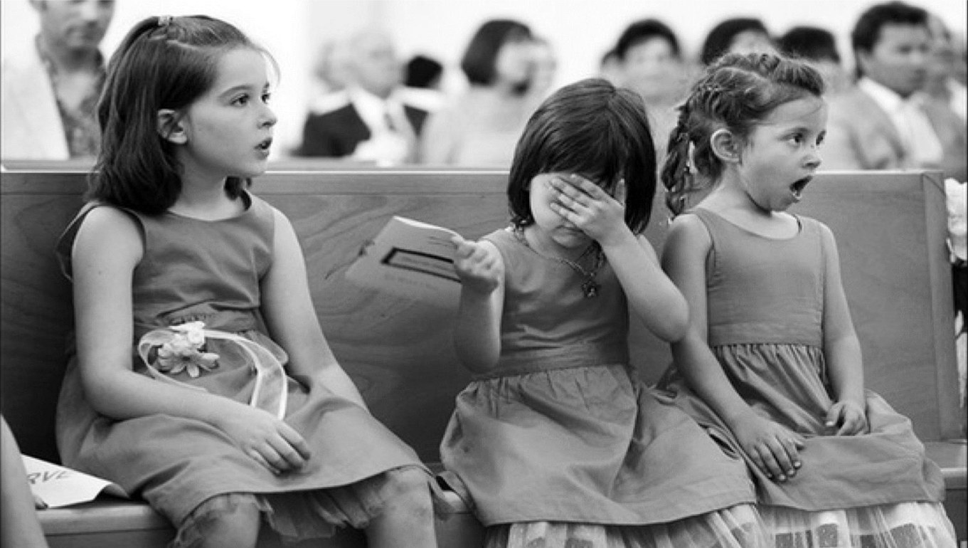 Black-and-white photo of three young girls in a church pew at a wedding. The girl on the left watches attentively, the girl in the middle covers her eyes, and the girl on the right gasps with her mouth wide open.